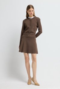 Abito in maglia marrone con scollatura rotonda, apertura con bottoni, dettagli con tasche e vita aderente. La gonna si allarga. Indossato con tacchi intrecciati color sabbia.