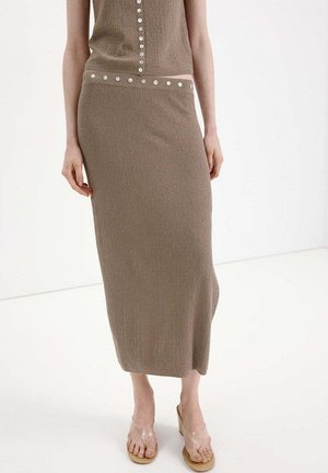 Vestido taupe sin mangas con detalles de botones en la parte superior y la cintura, combinado con sandalias de tacón transparentes.