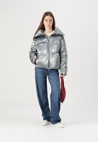 Casaco puffer prateado com gola alta, fecho de correr e bolsos, combinado com calças jeans de perna larga azuis claras e ténis brancos. A segurar uma bolsa vermelha.