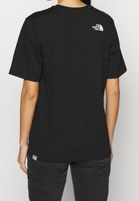Persona que lleva una camiseta negra de The North Face, de espaldas, combinada con jeans oscuros frente a un fondo claro y sencillo.