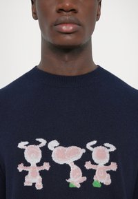 Navy blue knit sweater featuring a graphic design of three cartoon figures in pink with green accents, round faces, and distinct horns.