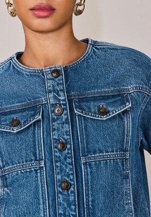 Žena v modrej rifľovej košeli na gombíky s dvoma vreckami na prsiach na gombíky a so zlatými kruhovými náušnicami.