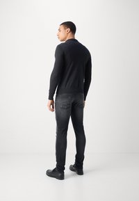 Mørk langærmet polo-shirt, tætsiddende design, båret med slidte sorte jeans. Modellen står med ansigtet vendt til venstre og fremviser ribbede ærmer og kant. Sorte sko.