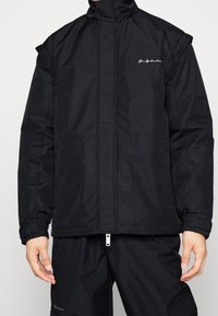 Svart vadderad jacka med hög krage, framsida med dragkedja och elastiska ärmslut. Har en broderad logotyp på bröstet och en slät textur.