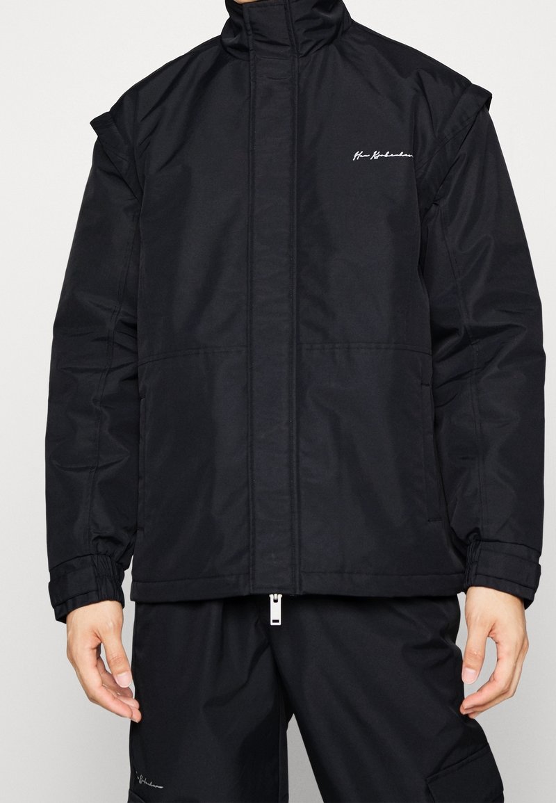 Svart vadderad jacka med hög krage, framsida med dragkedja och elastiska ärmslut. Har en broderad logotyp på bröstet och en slät textur.