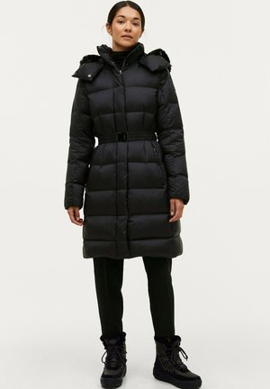 Zwarte pufferjas met een gezekerde taille, capuchon met nepbont afgewerkt, getailleerd ontwerp, zijzakken en een knielange snit. Draag het over zwarte broek en laarzen.