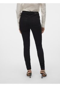 Femme portant un jean noir slim et des sandales à talons hauts noires, vue de dos sur un fond blanc uni.