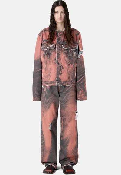 Jeune personne aux longs cheveux foncés, portant une veste et un pantalon assortis oversize délavés rouge et noir, usés, avec des chaussures marron, debout.