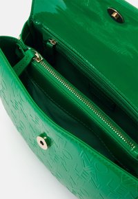 Grön lackläderväska med präglad text, gulddetaljer och inre dragkedjeficka. Har en jämn yta och en strukturerad design.