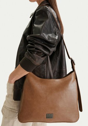 Osoba nosící černou koženou bundu a světlé kalhoty, nesoucí velkou hnědou koženou kabelku s nastavitelným popruhem.