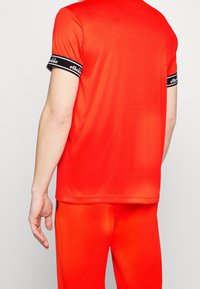 Rød atletisk T-shirt med korte ærmer og sort ribbet manchet med et logo. Stoffet er glat og strækbart, designet til aktivt brug.