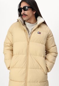 Beige pufferjack met een hoge kraag, voorzien van een voorrits en horizontale stiksels, vergezeld van een klein logo-opdruk op de borst.
