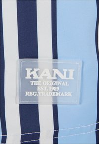 Mėlynos ir baltos juostelės audinys su aiškiu plastiko etiketu, kuriame užrašas "KANI ORIGINALAS EST. 1989 REG. PREKĖS ŽENKLAS" balta spalva.