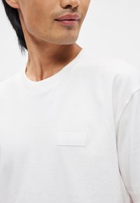 Λευκό βαμβακερό μπλουζάκι με στρογγυλή λαιμόκοψη και κοντά μανίκια. Χαρακτηριστική λεπτομέρεια είναι μια λευκή ορθογώνια ετικέτα στο στήθος.