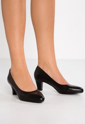 Sorte læderpumps med mellemhøj hæl, båret på bare fødder mod en lys baggrund, der viser siden og forsiden af skoene.