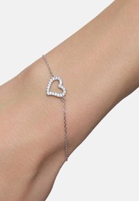 Srebrna bransoletka z zawieszką w kształcie serca, z przezroczystymi kamieniami wzdłuż brzegów. Łańcuszek jest delikatny i drobno powiązany.