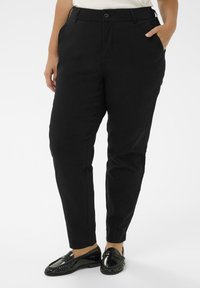 Persona, kas tērpusies melnās šaurās biksēs ar sānu kabatām, baltā augšpusē un melnās spīdīgās boršās, stāv pretī baltai fona.