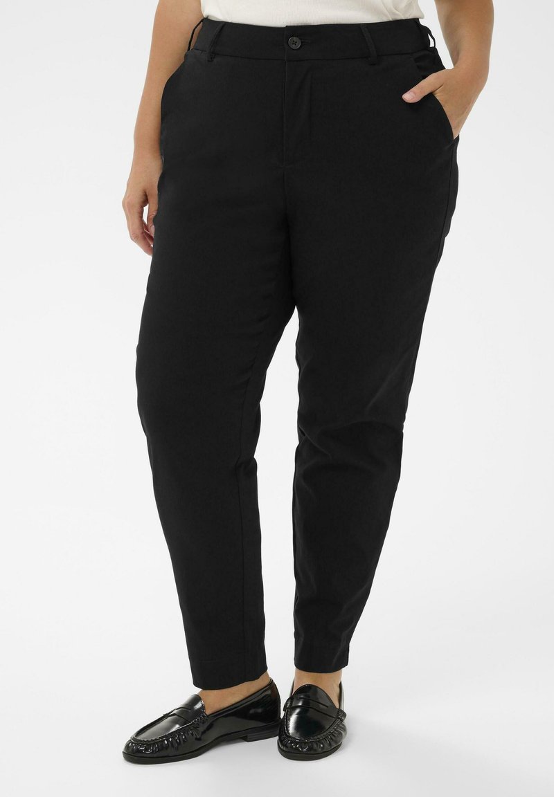 Persona, kas tērpusies melnās šaurās biksēs ar sānu kabatām, baltā augšpusē un melnās spīdīgās boršās, stāv pretī baltai fona.