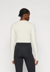 Lichtrompe crèmekleurig longsleeve shirt gemaakt van een glad materiaal, gecombineerd met zwarte high-waisted leggings. Heeft een klein logo bij de nek.