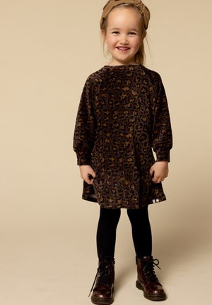 Een jong meisje dat lacht, gekleed in een jurk met luipaardprint, zwarte leggings, bruine veterschoenen en een beige hoofdband tegen een beige achtergrond.