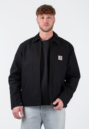 Black jacket with a brown corduroy collar, front zipper, left chest pocket, and branded logo. Worn over a black t-shirt and light blue jeans.