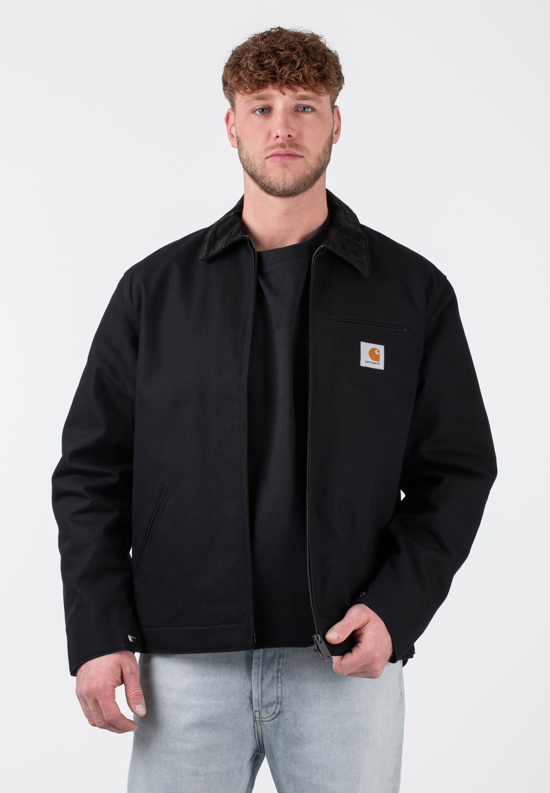 Schwarze Jacke mit einem braunen Cordkragen, Frontreißverschluss, linker Brusttasche und markiertem Logo. Getragen über einem schwarzen T-Shirt und hellblauen Jeans.