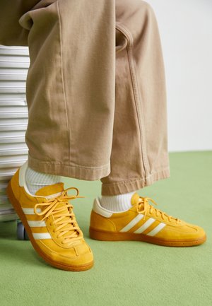 Persona con pantalones beige y calcetines blancos sentada, mostrando zapatillas Adidas amarillas brillantes con rayas blancas sobre suelo verde.