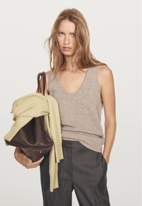 Žena se světle hnědými vlasy má na sobě šedý bezrukávový top a tmavé kalhoty, drží velkou hnědou tašku, na které je přehozený světlounce zelený svetr.