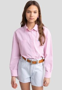 Camicia button-up a righe rosa chiaro con maniche lunghe, una tasca singola e un piccolo logo blu sul petto. Abbinata a pantaloni corti di denim chiaro e una cintura marrone.