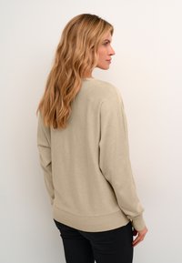 Beige pulóver bordás mandzsettával és alsó szegéllyel, puha anyagból készült. Laza szabású és kerek nyakkivágása van, sötét nadrággal viselve.