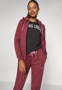 Maroon kapuutsiga dressipluus, millel on heledat roosat nööri, koos tumehallide särgi ja vastavate pükstega. Omab logo aktsente. Pehme, juhuslik disain.