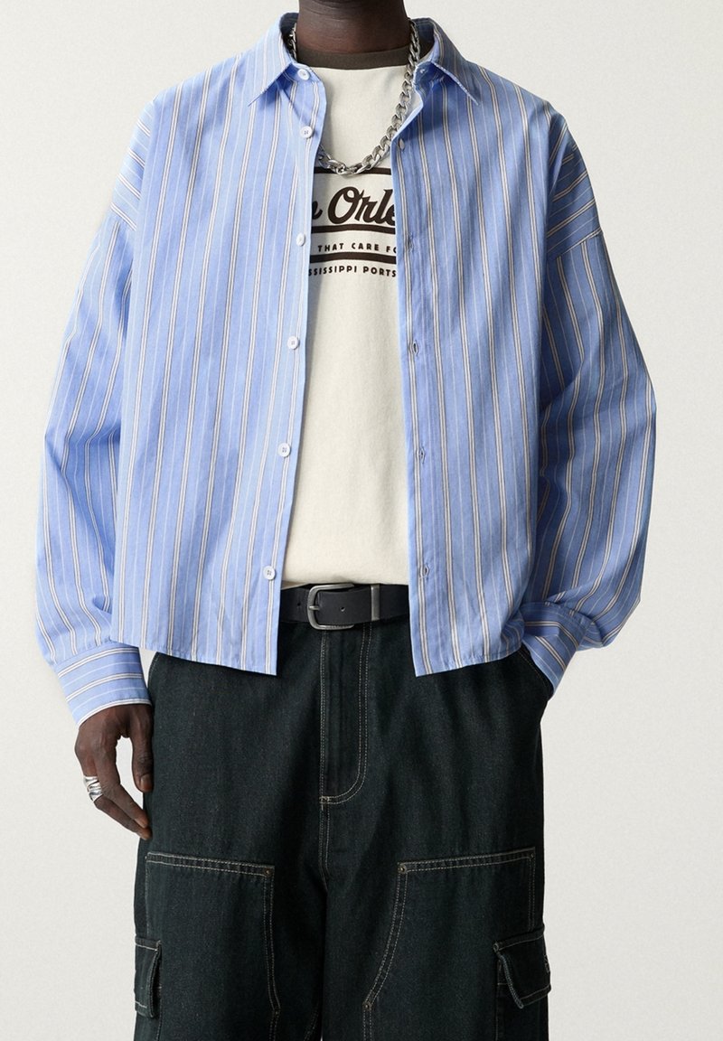 Chemise à rayures bleues et blanches à boutons, à manches longues, portée avec un t-shirt de couleur claire et un pantalon cargo foncé. Collier chaîne argentée visible.