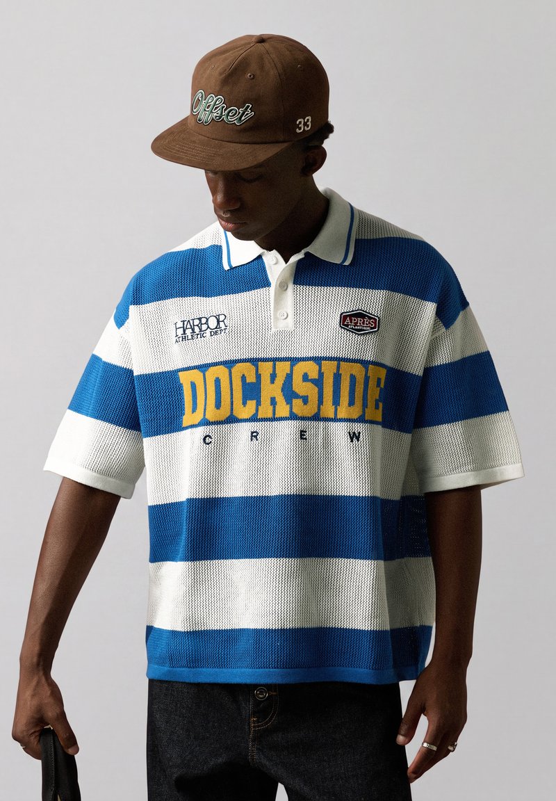 Homme portant une casquette marron et un polo rayé bleu et blanc avec un texte jaune "DOCKSIDE", regardant vers le bas sur un fond uni.