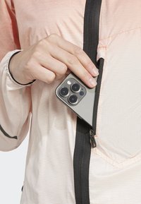 Veste légère de couleur pêche avec une poche zippée. Un smartphone gris est en train d'être placé dans la poche. Tissu texturé avec un motif en quadrillage.