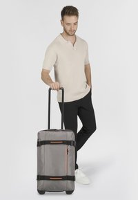 Maleta de ruedas gris con una textura de tela suave, que presenta correas negras, un acento naranja y un bolsillo lateral con cremallera.