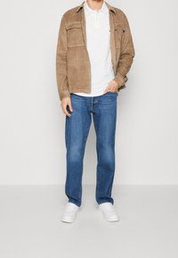 Brun kordfløyelsjacka över vit polotröja, tillsammans med blå denimjeans och vita sneakers. Enkel, avslappnad design med fickor.