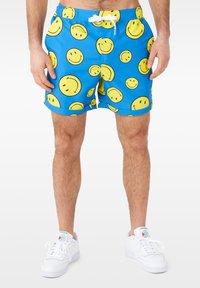 Shorts de bain bleus avec un motif de visages souriants jaunes, taille élastique avec cordon de serrage, et baskets blanches.