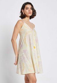Ärmelloses gelbes Kleid mit lockerer Passform, das lila Blumenmuster und Schriftzüge aufweist. Hergestellt aus weichem Material, hat es einen V-Ausschnitt und dünne Träger.