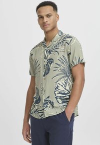 Chemise à manches courtes avec une base verte ornée de grands motifs de feuilles tropicales bleu marine, fermeture à boutons et coupe décontractée.