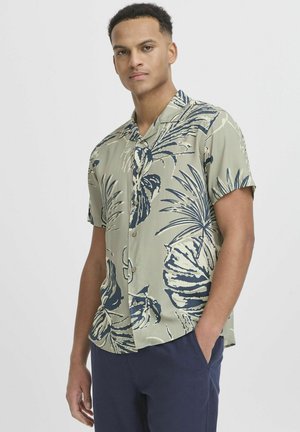 Cămașă cu mânecă scurtă, cu o bază verde și modele mari de frunze tropicale bleumarin, cu nasturi în față și o croială lejeră.