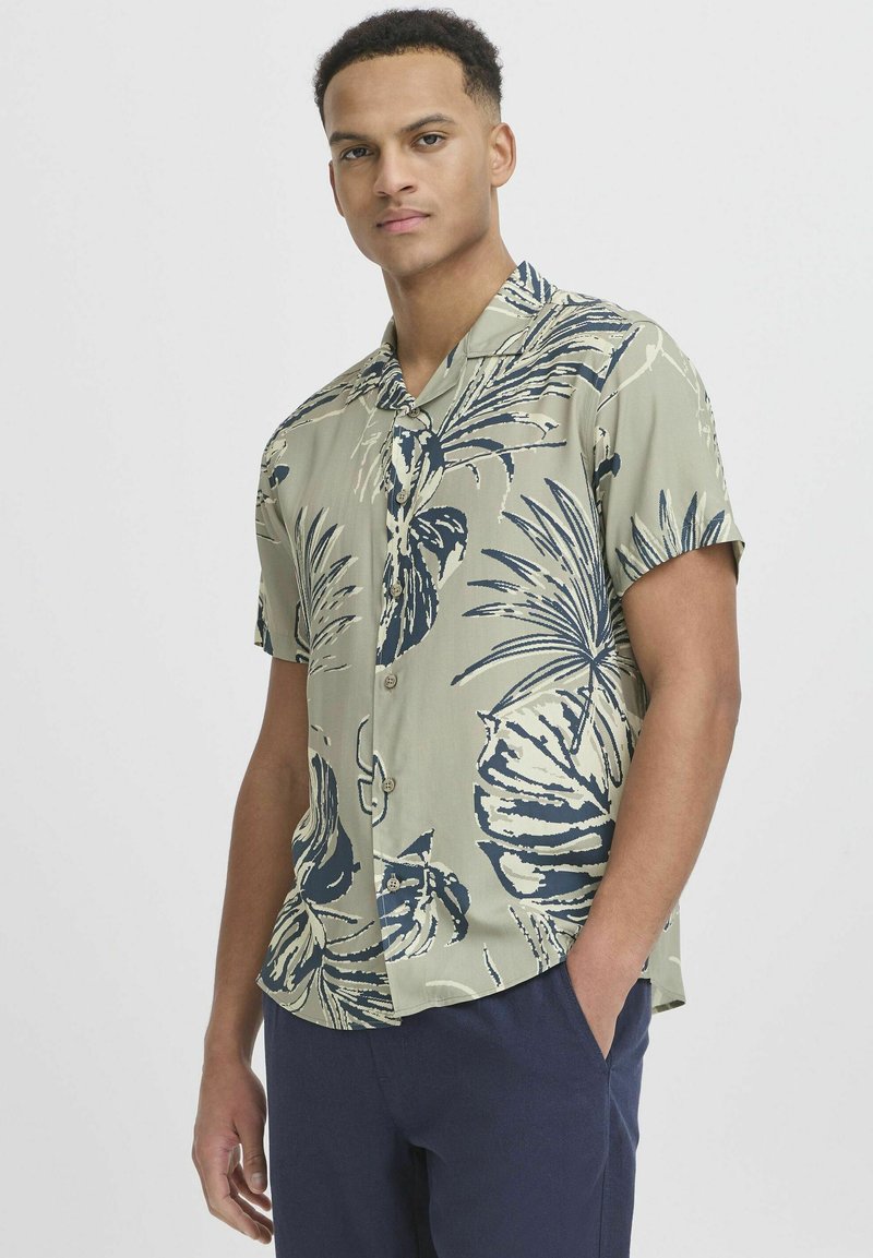 Chemise à manches courtes avec une base verte ornée de grands motifs de feuilles tropicales bleu marine, fermeture à boutons et coupe décontractée.