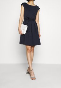 Žena v modro-černé šaty ke kolenům s uvázaným pasem, držící bílou kabelku a nazouvající černo-bílé pruhované sandály na podpatku.