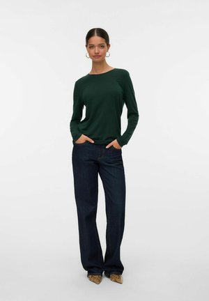 Vrouw met donker haar in een groene blouse met lange mouwen, donkere spijkerbroek met wijde pijpen en bruine puntige schoenen, staand met de handen in de zakken op een witte achtergrond.