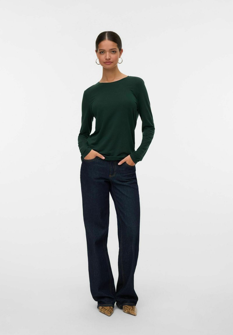 Frau mit dunklem Haar, in einem grünen Langarmshirt, dunkelblauen weit geschnittenen Jeans und braunen spitzen Schuhen, die mit den Händen in den Taschen vor einem weißen Hintergrund steht.