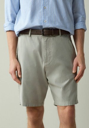 Mann trägt hellgraue, strukturierte Shorts mit einem braunen Ledergürtel und ein blau-weiß gestreiftes Hemd mit Knöpfen und hochgekrempelten Ärmeln.