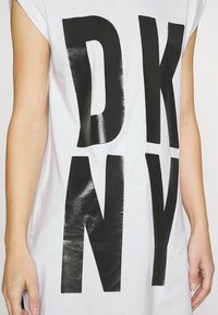 Baltas trumpas rankovėmis marškinėliai su dideliu, blizgiu juodu "DKNY" spaudiniu priekyje. Minkšta medvilnės medžiaga, laisvas siluetas ir apvalus kaklas.