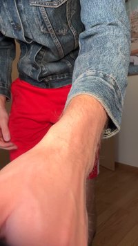 Džínová bunda s klasickým strihom, vystavujúca modrú textúru a viditeľné švy, spárovaná s červenými šortkami. Ruka sa predlžuje dopredu, ruka sa siaha.