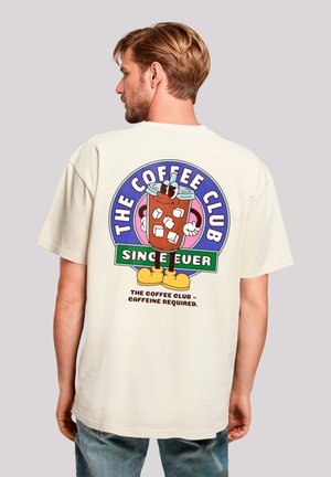 Cream-colored t-shirt featuring a playful graphic of a drink with a straw, ice cubes, and the text "THE COFFEE CLUB" and "Caffeine required."