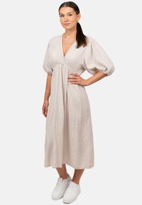 Lichtbeige midi-jurk met een diepe V-hals, pofmouwen, gestructureerde stof en een geraffineerde taille; gecombineerd met witte sneakers.