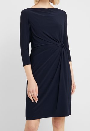 Woman wearing a navy blue knee-length dress with three-quarter sleeves and a twisted knot detail at the waist.